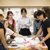Students sit and stand around a table while a professor looks down at their work.